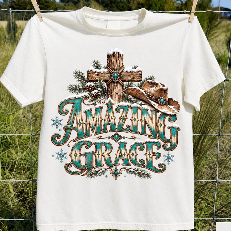 May include: A cream-coloured short-sleeve t-shirt featuring a graphic print of a wooden cross covered in snow, a brown cowboy hat, and pine branches. The text "AMAZING GRACE" is printed in large, decorative teal and brown lettering with snowflakes.