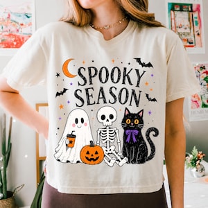 May include: A cream-colored t-shirt featuring a graphic print with the text SPOOKY SEASON in black letters. The design includes a white ghost holding an orange cup, a skeleton, a jack-o'-lantern, a black cat with a purple bow, and bats.