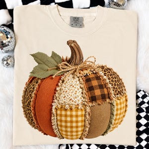May include: A cream-colored short-sleeve t-shirt featuring a large graphic of a patchwork pumpkin. The pumpkin is composed of various fabric patterns including leopard print, orange solid, floral, brown plaid, and yellow gingham with frayed edges.