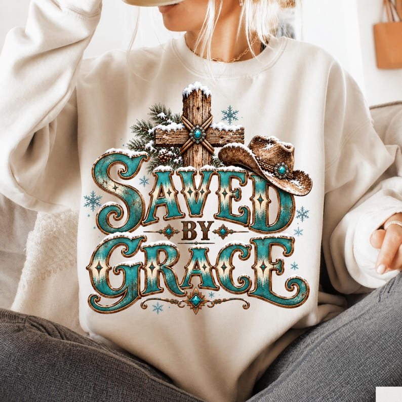 May include: A cream-coloured crewneck sweatshirt featuring a graphic print of a wooden cross covered in snow, a brown cowboy hat, and the text "SAVED BY GRACE" in large, turquoise, western-style lettering with snowflake accents throughout the design.
