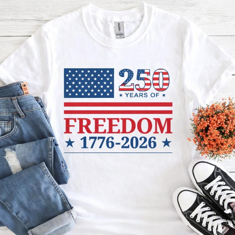 May include: White t-shirt with a patriotic design. The design features a blue rectangle with white stars, red and white stripes, and the words "250 YEARS OF FREEDOM 1776-2026" in red and blue. The shirt is paired with blue jeans and black sneakers.