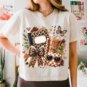 May include: A cream-colored t-shirt featuring a graphic print of a leopard-print notebook, a bow, a pencil, an iced coffee, and an apple wearing heart-shaped sunglasses. The design includes a butterfly, small hearts, and star accents in brown tones.