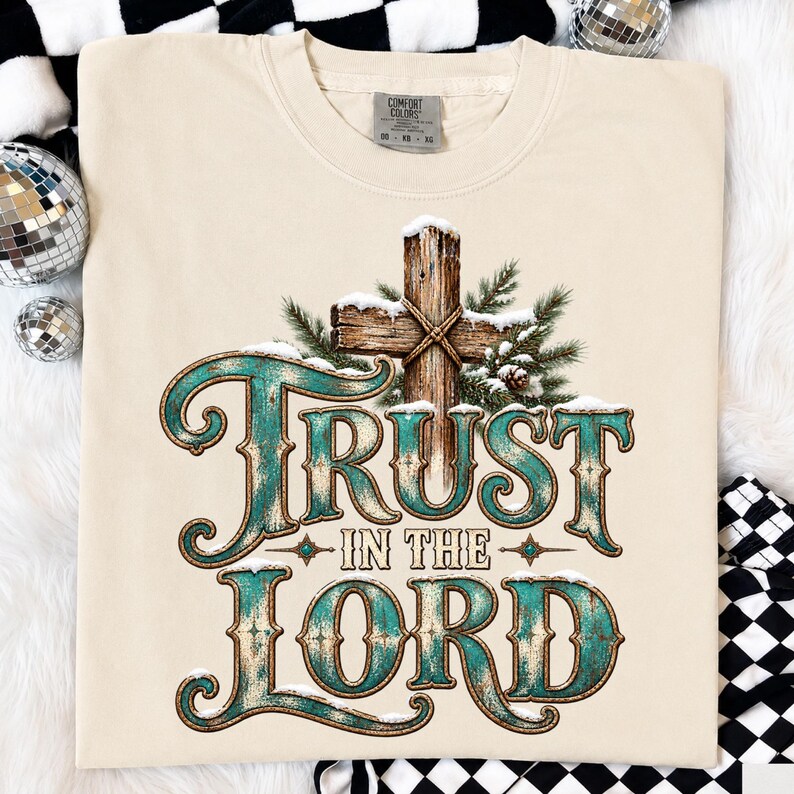May include: A beige short-sleeve t-shirt featuring a graphic of a wooden cross covered in snow and pine branches. The text "TRUST IN THE LORD" is printed in a large, distressed teal font with gold outlines. The shirt has a Comfort Colors brand label.