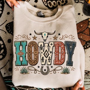 May include: A beige crewneck sweatshirt featuring a graphic print of a steer skull above the word HOWDY in large, patterned block letters. The design includes decorative stars, a horseshoe, rope elements, and small agave plants in earthy tones.