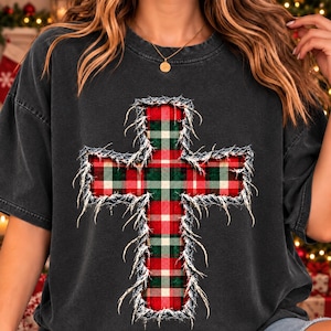 May include: A charcoal gray short-sleeve t-shirt featuring a large, central cross graphic. The cross has a red, green, and white plaid pattern with a distressed, frayed-edge effect around its entire perimeter, giving it a textured appearance.