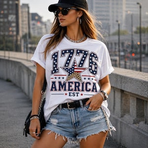 May include: White t-shirt with a patriotic design featuring the year "1776" in blue and white stars, a star with a leopard print pattern, and the word "AMERICA" in red and blue. The shirt is paired with denim shorts and a black belt.