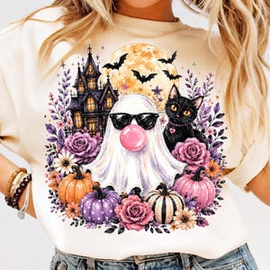 May include: A cream-colored t-shirt featuring a graphic print of a ghost wearing black sunglasses and blowing a pink bubble, a black cat, a haunted house, a full moon with bats, purple roses, and orange, purple, and striped pumpkins.