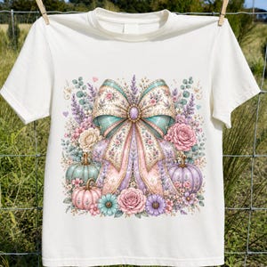 May include: A white short-sleeve t-shirt featuring a central graphic of a large, ornate bow in pastel shades of pink, blue, and purple, surrounded by detailed illustrations of roses, lavender sprigs, eucalyptus leaves, and small pumpkins.