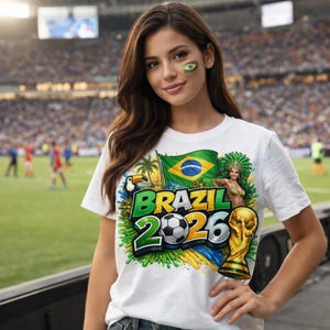 May include: White t-shirt with a colorful graphic design featuring the Brazilian flag, a toucan, a woman in a carnival costume, a soccer ball, and the text "BRAZIL 2026". A gold trophy is also visible.