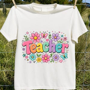 May include: A white short-sleeved t-shirt featuring a colorful graphic on the chest. The word Teacher is printed in large, multi-colored bubble letters, surrounded by various illustrations of flowers, small hearts, stars, and two butterflies.