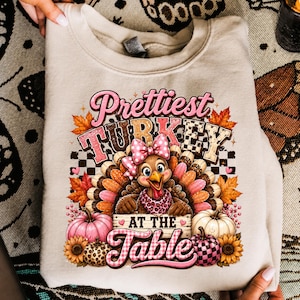 May include: A beige crewneck sweatshirt featuring a graphic of a turkey wearing a pink polka-dot bow and a leopard-print scarf. The text reads "Prettiest TURKEY AT THE TABLE" surrounded by autumn leaves, pink pumpkins, and sunflowers.