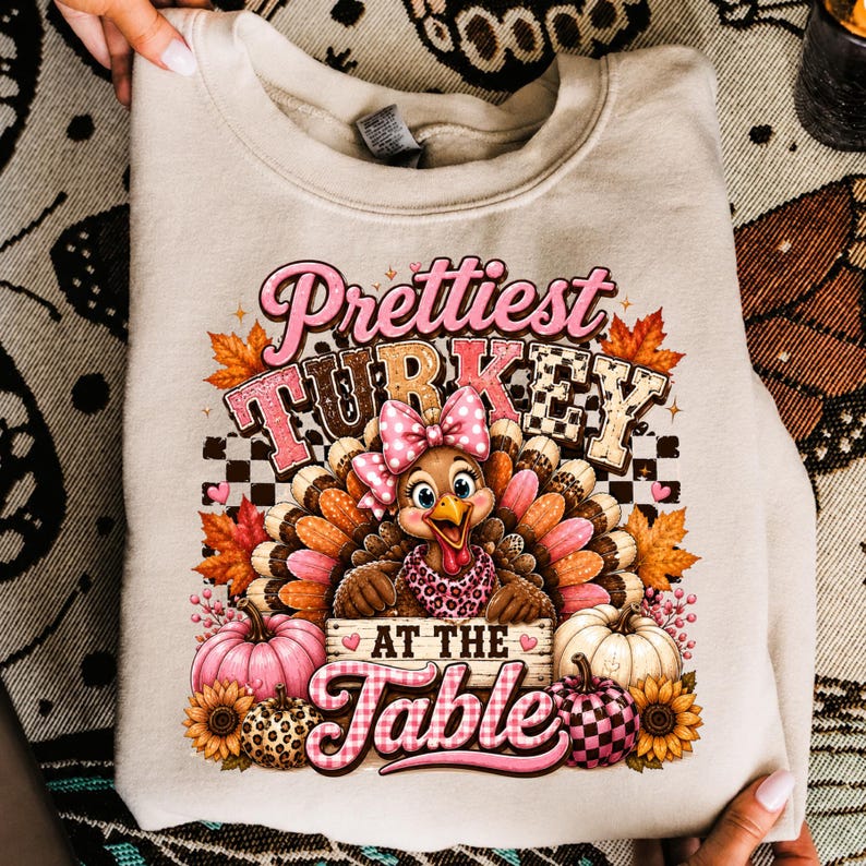 May include: A beige crewneck sweatshirt featuring a graphic of a turkey wearing a pink polka-dot bow and a leopard-print scarf. The text reads "Prettiest TURKEY AT THE TABLE" surrounded by autumn leaves, pink pumpkins, and sunflowers.