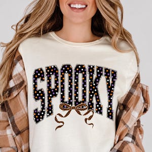 May include: A cream-coloured crew neck t-shirt featuring the word SPOOKY in large block letters. The letters are black with a pattern of white, orange, and purple polka dots. Below the text is a brown leopard print bow graphic.