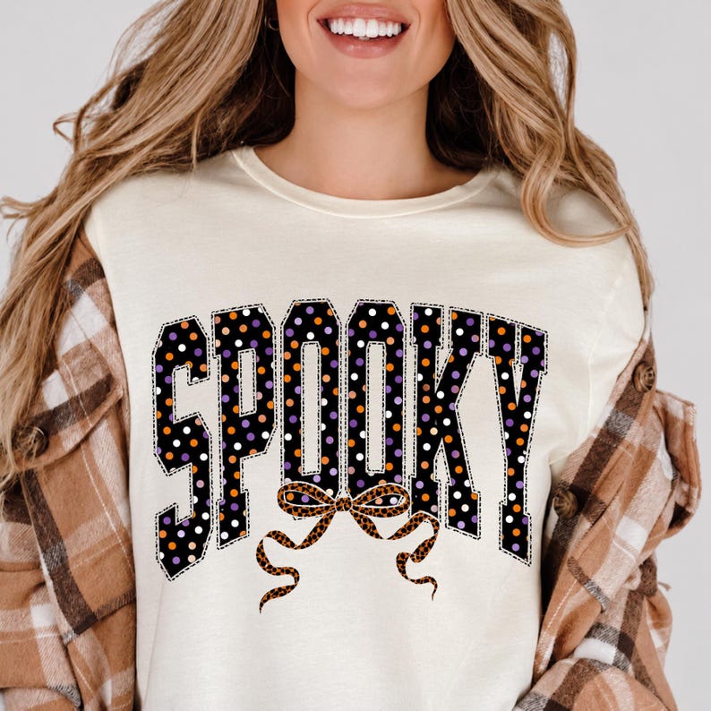 May include: A cream-coloured crew neck t-shirt featuring the word SPOOKY in large block letters. The letters are black with a pattern of white, orange, and purple polka dots. Below the text is a brown leopard print bow graphic.