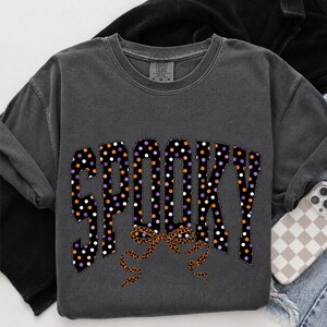 May include: A charcoal grey crewneck t-shirt featuring the word SPOOKY in large block letters. The letters are black with a pattern of white, orange, and purple polka dots. Below the text is a leopard print bow graphic with long ribbons.