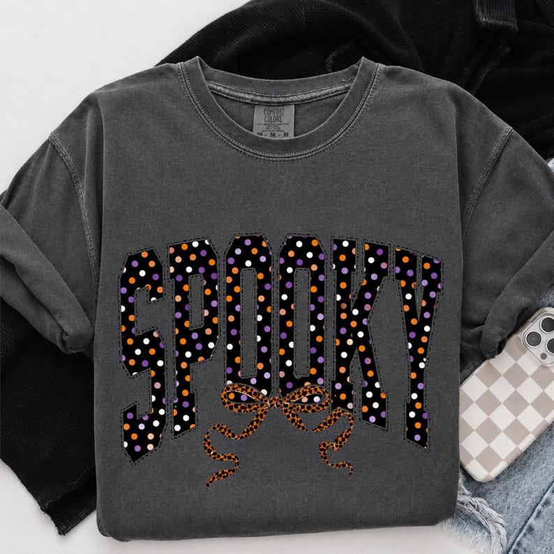 May include: A charcoal grey crewneck t-shirt featuring the word SPOOKY in large block letters. The letters are black with a pattern of white, orange, and purple polka dots. Below the text is a leopard print bow graphic with long ribbons.