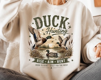 May include: A cream-colored crewneck sweatshirt featuring a graphic print of three ducks in a marshy landscape. The text reads DUCK Hunting, RISE AIM HUNT, and BORN TO HUNT BUILT FOR THE WILD. The design includes cattails, a sun, and trees.