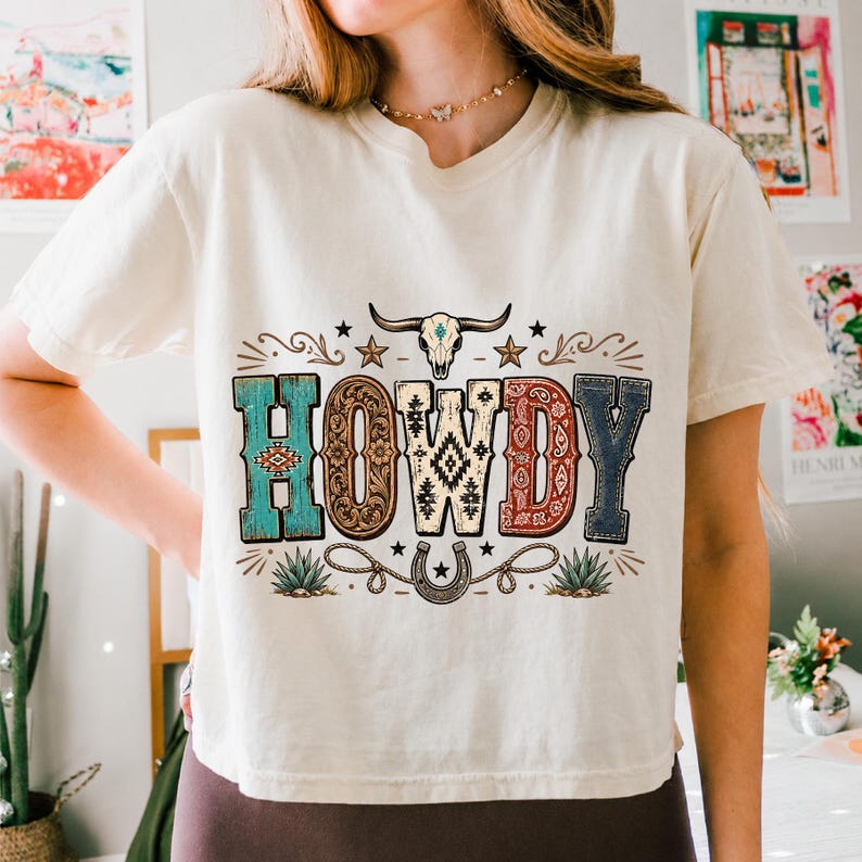 May include: A cream-colored short-sleeve t-shirt featuring a graphic print with the word HOWDY in large, patterned letters. The design includes a steer skull, stars, a horseshoe with a rope, and agave plants in shades of turquoise, brown, and red.
