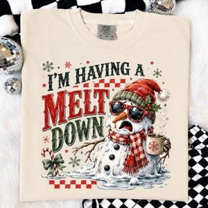 Peut inclure: Un t-shirt couleur cr&egrave;me avec l'illustration d'un bonhomme de neige en train de fondre, portant un bonnet rouge, des lunettes de soleil rouges et une &eacute;charpe &agrave; carreaux rouges et blancs. Il tient une tasse fumante. Texte : I'M HAVING A MELT DOWN.