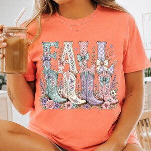 May include: A coral-colored t-shirt featuring the word FALL in large letters, each decorated with floral patterns and bows. Below the letters are four cowboy boots in shades of teal, white, purple, and pink, surrounded by various colorful flowers.