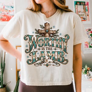 May include: A cream-coloured short-sleeve t-shirt featuring a graphic print of a wooden cross covered in snow and pine branches, with the text Worthy Is The Lamb in teal and brown lettering, accompanied by a small white lamb and blue snowflakes.
