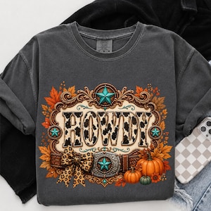 May include: A charcoal gray crewneck t-shirt featuring a graphic print of a brown western-style belt buckle frame with the text HOWDY in a cow print pattern, accented by orange autumn leaves, a leopard print bow, and orange pumpkins.