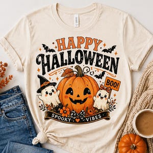 May include: Cream-colored t-shirt with a Halloween-themed graphic. The design features a smiling jack-o'-lantern, two ghosts, bats, and the words "Happy Halloween" and "Spooky Vibes." A pair of blue jeans and a cup of coffee are next to the shirt.
