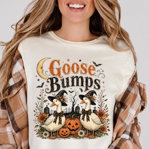 May include: A cream-colored t-shirt featuring a graphic print with the text "Goose Bumps" in orange and black. The design includes two white geese wearing black witch hats and polka-dot bows, surrounded by pumpkins, sunflowers, and a crescent moon.