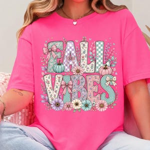 May include: A bright pink t-shirt featuring the text FALL VIBES in large, blocky letters. The letters are decorated with floral patterns, checkered prints, and polka dots, accented by illustrations of pumpkins, bows, and various colorful flowers.
