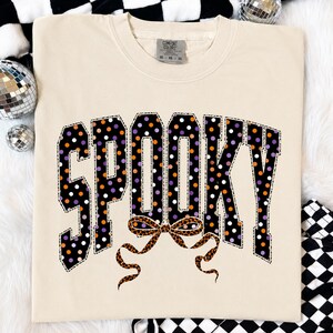 May include: A cream-coloured short-sleeve t-shirt featuring the word SPOOKY in large, block-style letters. The letters are black with a polka dot pattern in orange, purple, and white. A leopard print bow is positioned below the text.