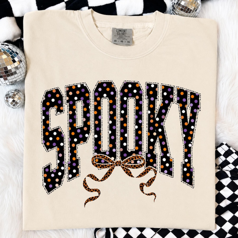 May include: A cream-coloured short-sleeve t-shirt featuring the word SPOOKY in large, block-style letters. The letters are black with a polka dot pattern in orange, purple, and white. A leopard print bow is positioned below the text.