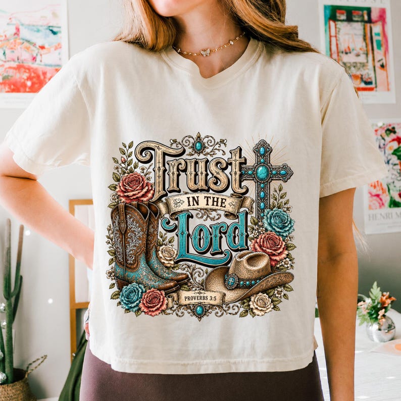 May include: A cream-coloured t-shirt featuring a graphic print with the text "Trust in the Lord" and "Proverbs 3:5". The design includes a turquoise and brown cowboy boot, a brown cowboy hat, a turquoise cross, and various colourful roses.
