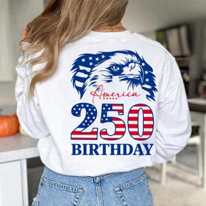 May include: White long-sleeve shirt with a patriotic design. Features a blue and white eagle graphic, the word "America" in red script, and "250 BIRTHDAY" in red and blue. The shirt is made of a soft material.