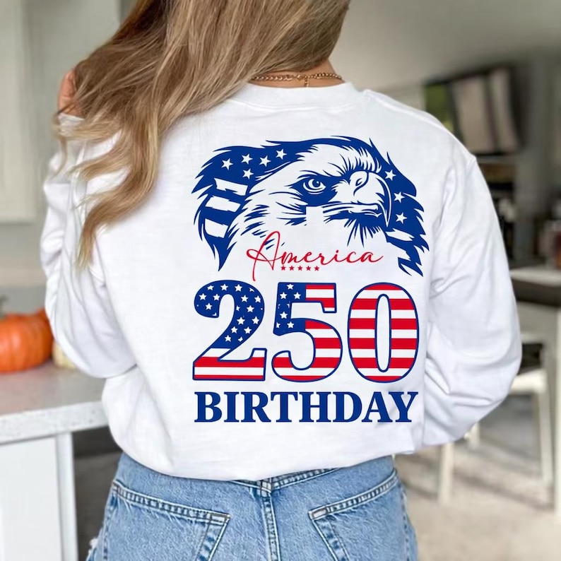 May include: White long-sleeve shirt with a patriotic design. Features a blue and white eagle graphic, the word "America" in red script, and "250 BIRTHDAY" in red and blue. The shirt is made of a soft material.