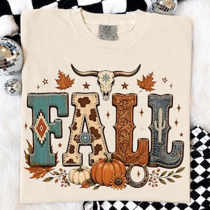 May include: A cream-colored t-shirt featuring the word FALL in large decorative letters. The F is teal with a geometric pattern, the A is cow print with a turquoise concho, the L is brown floral, and the L is a blue cowboy boot. A bull skull, pumpkins, and leaves are included.