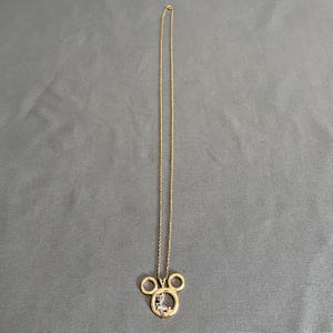 May include: A gold-colored necklace with a pendant shaped like Mickey Mouse's head. The pendant has a small figure inside and is suspended from a delicate chain. The necklace is displayed against a gray background.