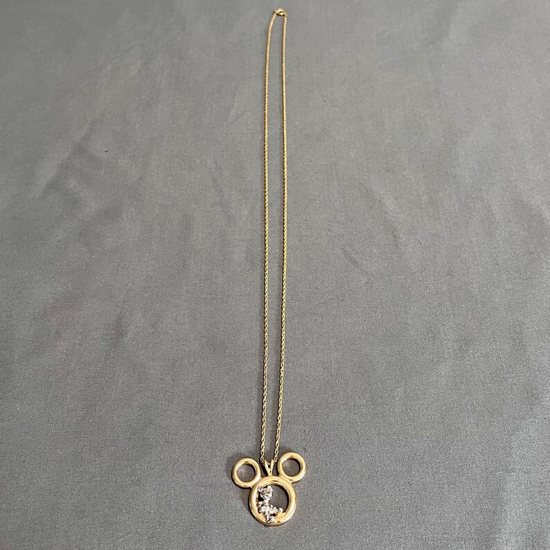 May include: A gold-colored necklace with a pendant shaped like Mickey Mouse's head. The pendant has a small figure inside and is suspended from a delicate chain. The necklace is displayed against a gray background.