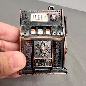 Puede incluir: Una hucha de metal en miniatura con forma de máquina tragaperras, acabado en bronce oscuro y reflejos cobrizos. El frente presenta un emblema de águila, el texto GOLDEN EAGLE, una ranura para monedas de 10 centavos y una tabla de premios.