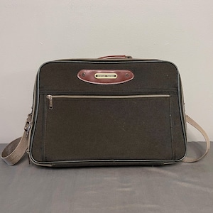 May include: A dark olive green rectangular travel bag with a brown leather accent and the text "American Tourister". The bag has a zippered front pocket, a top handle, and a tan shoulder strap.