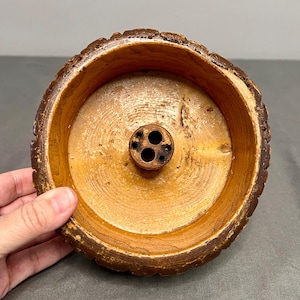 May include: A wooden incense holder with a natural, rustic aesthetic. The holder is circular, featuring a light brown interior and a darker, textured rim resembling tree bark. It has a central area with three holes for incense sticks.