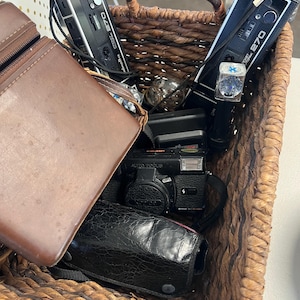 May include: A woven basket filled with vintage cameras and accessories. The basket contains several black Minolta 270 cameras, a black camera with a leather case, and a brown leather camera case. The cameras are in various conditions, suggesting they are used or collectible.