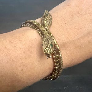 Peut inclure: Un bracelet en métal doré en forme de serpent enroulé avec des écailles texturées couvrant toute la longueur du corps. Le design présente deux têtes de serpent distinctes aux extrémités, chacune détaillée avec de petits accents verts pour les yeux.