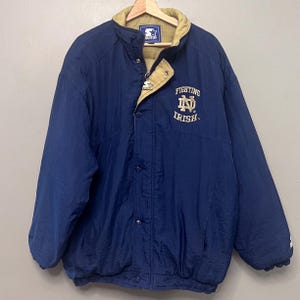 Puede incluir: Una chaqueta azul marino de la marca Starter con forro y cuello de color beige. La parte delantera muestra el texto bordado Fighting Irish con un logotipo dorado. Incluye una cremallera completa, cierre de botones a presión y tirador Starter.