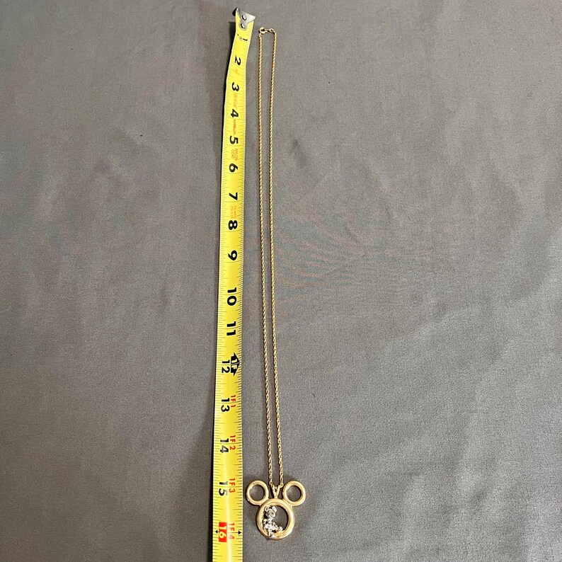 May include: A gold-colored necklace with a Mickey Mouse pendant. The pendant is approximately 1.5 inches wide and 1.25 inches tall. The chain is about 15 inches long. The pendant has a clear, sparkly design inside the Mickey Mouse head.