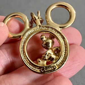 May include: A gold-toned Mickey Mouse pendant. The pendant features Mickey Mouse inside a circular frame, with two circular ears above. The word "Disney" is inscribed at the bottom of the frame. The pendant is attached to a chain.