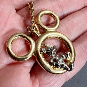 May include: Gold-tone pendant necklace featuring a Mickey Mouse design. The pendant has a gold-colored outline in the shape of Mickey's head, with a silver-colored Mickey Mouse figure inside. The necklace chain is also gold-colored.