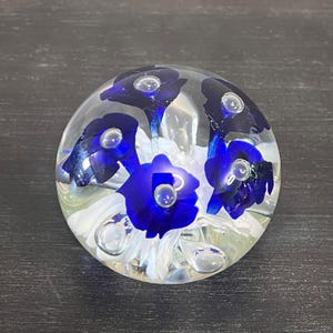 May include: A spherical glass paperweight featuring a clear outer layer encasing deep blue floral shapes and white petal-like structures. Small air bubbles are suspended within the glass, creating a layered effect throughout the interior design.