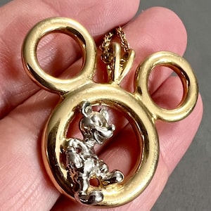 May include: Gold-tone Mickey Mouse pendant necklace. The pendant features a gold outline of Mickey Mouse's head with a silver-colored Mickey Mouse figure inside. The necklace has a delicate gold chain.