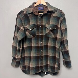 Puede incluir: Una camisa de manga larga con botones y un patrón de cuadros en tonos marrón oscuro, verde azulado y bronceado. La camisa incluye un cuello puntiagudo, dos bolsillos en el pecho con solapas puntiagudas y botones de presión metálicos plateados.