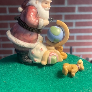 May include: A ceramic Santa Claus figurine in a red suit and hat, examining a globe. A small dog figurine and a sack of gifts are also present. The figurines are on a green surface.